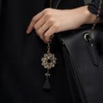 Elegant black handbag adorned with a floral keychain, featuring a gold and crystal design, held by a woman in a stylish outfit.