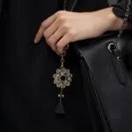Elegant black handbag adorned with a floral keychain, featuring a gold and crystal design, held by a woman in a stylish outfit.