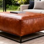Stylish brown leather ottoman with a sleek metal frame, placed in a modern living room setting, complemented by greenery and a cozy sofa.