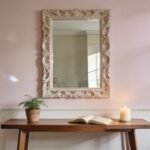 Elegant interior scene featuring a decorative mirror, wooden table, potted plant, open book, and a lit candle against a soft pink wall.