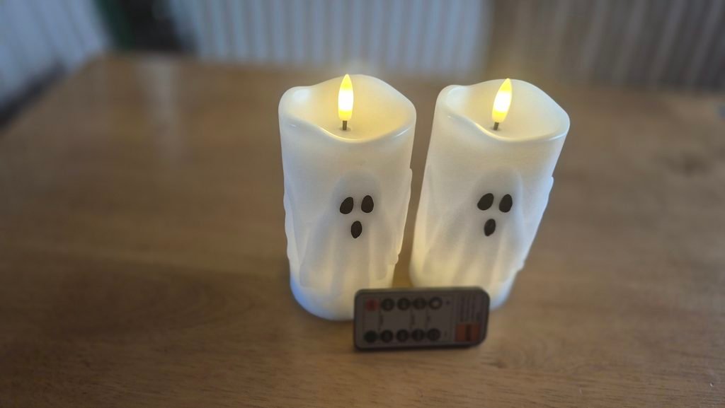 Two ghost-themed flameless pillar candles with remote on wooden table, perfect for creating spooky Halloween vibes.